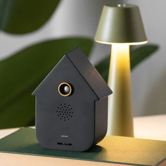 Zwarte Karlsson Modern Cuckoo sfeerbox decoratief geplaatst op een boek naast een brandende lamp
