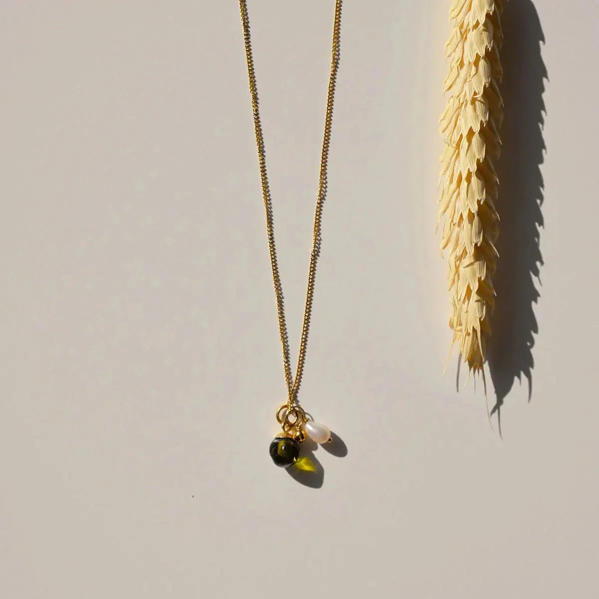 Sfeerfoto van de goudkleurige Vinoos ketting Ode to Green Stilte met groene glaskraal naast een droogbloem