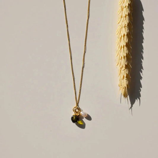 Sfeerfoto van de goudkleurige Vinoos ketting Ode to Green Stilte met groene glaskraal naast een droogbloem